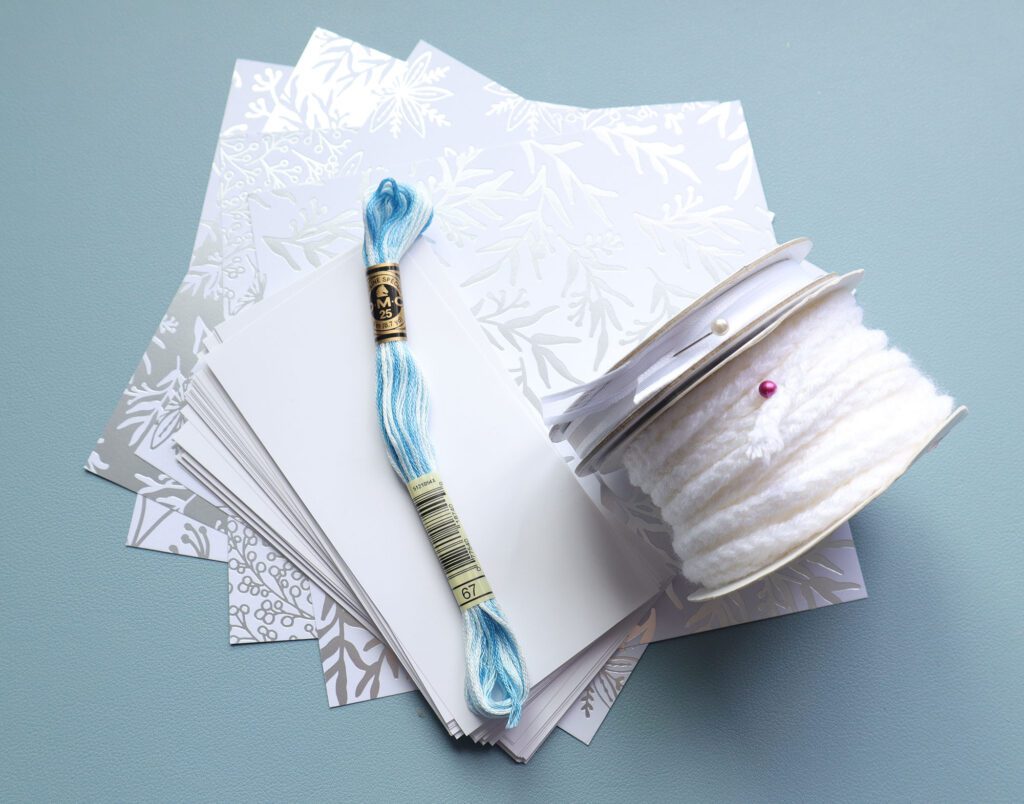paper, decorative cardstock, blue embroidery thread and two types of ribbon sitting on a blue backdrop to show what materials are needed for the mini chain stitch pamphlet tutorial