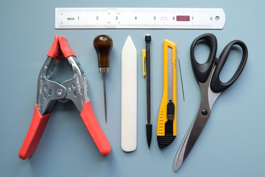 bookbinding tools clamp awl bone folder olfa knife shears ruler and pencil on a blue mat