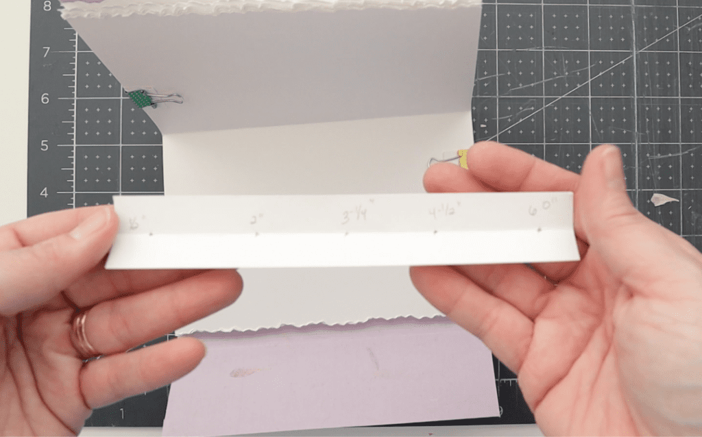 template strip with five punch holes and measurements marked using a pencil, held in two hands above a cutting surface