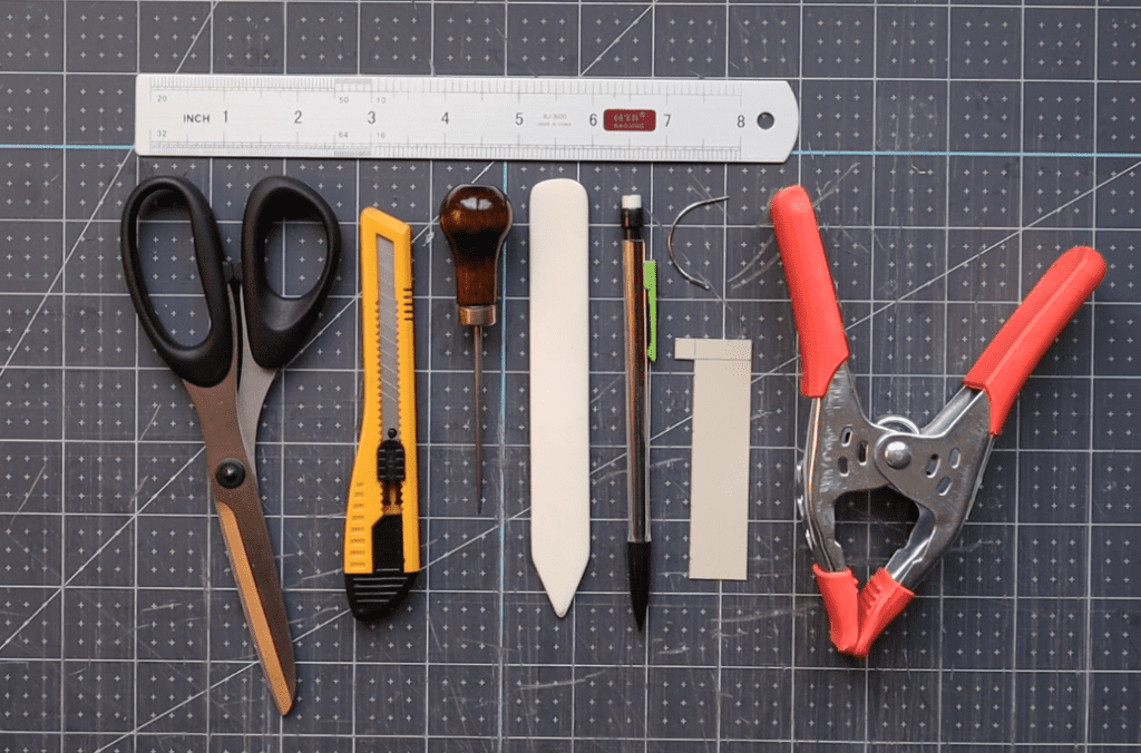tools needed to sew a coptic binding laying on a cutting mat, including scissors, bone folder, awl, clamp, ruler, olfa knife, pencil and template