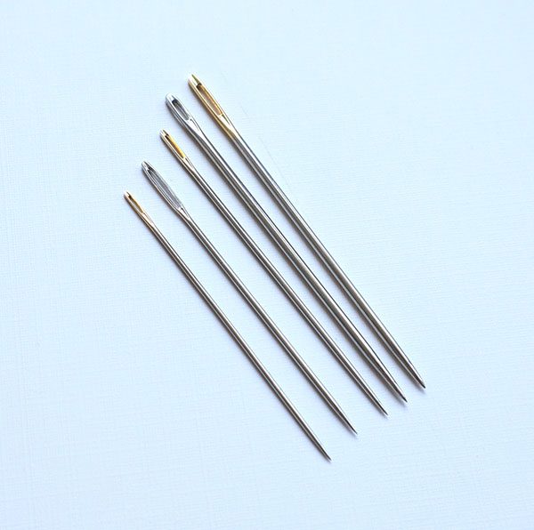 five different book binding making needles laying side by side on a blue backdrop